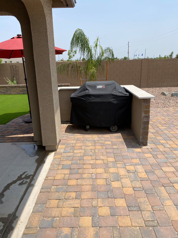  built-in outdoor BBQ station made of stone and brick, featuring a grill, countertop workspace, and storage cabinets. Surrounding the BBQ area are flagstone pavers and lush landscaping, creating an inviting outdoor cooking space